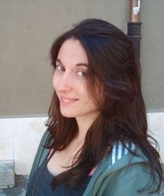 Elena Asensio. Una mujer con el pelo largo y oscuro y vestida con una camisa gris y una chaqueta verde claro está al aire libre. Ella sonríe levemente y mira hacia la cámara. El fondo presenta una pared beige y parte de la estructura de un edificio.