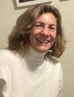 Elena Pol. Una mujer con el pelo hasta los hombros y un jersey de cuello alto de color claro sonríe cálidamente a la cámara. Está en el interior, con una pared de color claro y cuadros enmarcados parcialmente visibles al fondo.