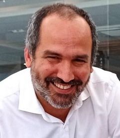 Abel Ponce. Un hombre de pelo corto oscuro, barba y bigote sonríe. Viste una camisa blanca y está fotografiado sobre un fondo con paneles de vidrio y un indicio de estructura de madera.