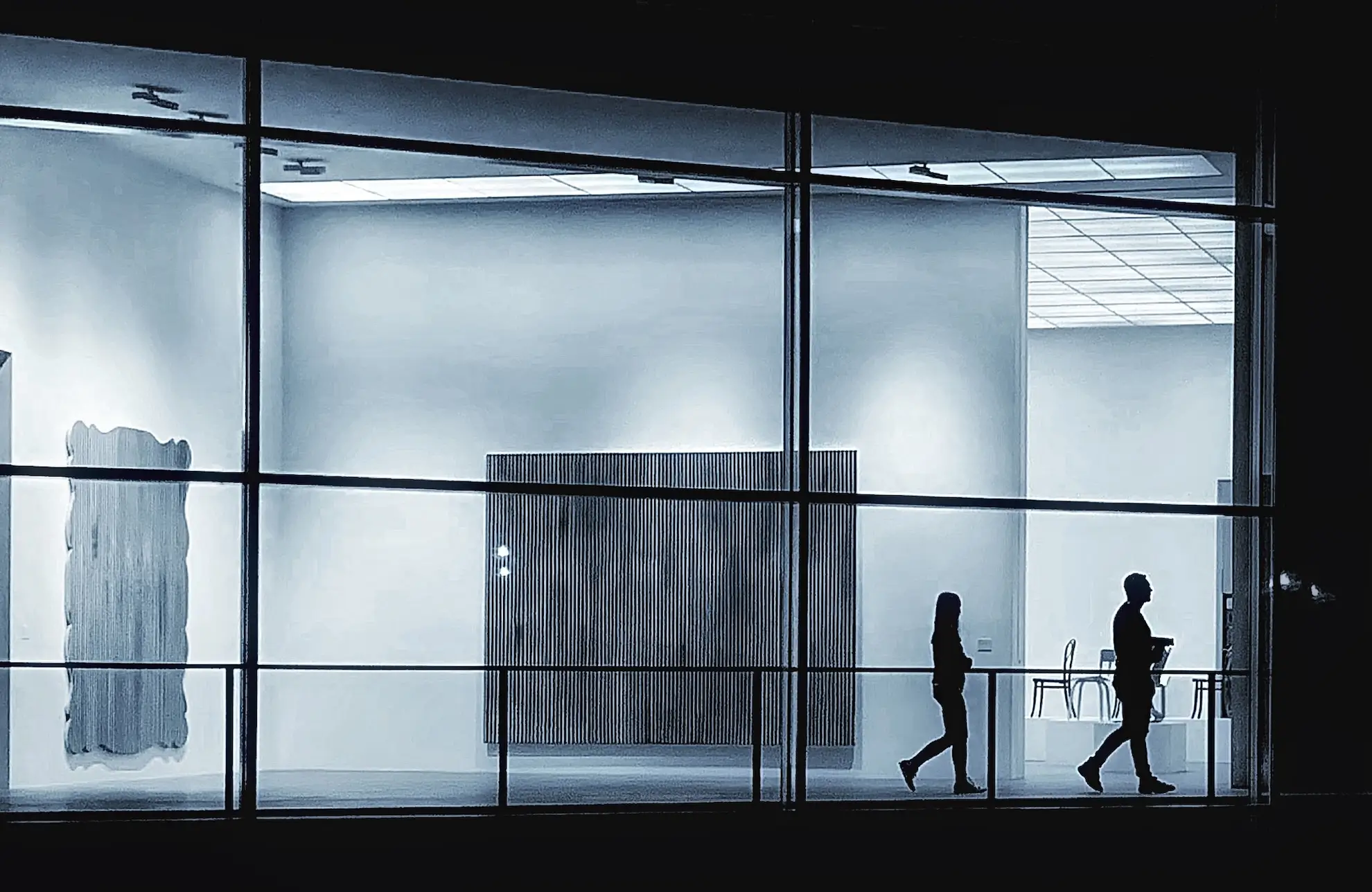 Una vista nocturna a través de una gran ventana de vidrio muestra dos siluetas de personas caminando en direcciones opuestas dentro de una galería u oficina moderna y bien iluminada. La habitación presenta una decoración minimalista, que incluye arte abstracto en las paredes y un panel de madera.