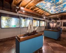 Una exposición de museo que presenta modelos de barcos detallados en cajas de vidrio sobre pedestales azules. La sala tiene un techo de madera, con mapas y obras de arte náutico que adornan las paredes y un techo pintado que representa una escena marítima.
