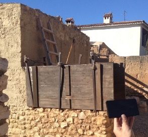 Una persona sostiene un teléfono inteligente frente a un edificio rústico de piedra en construcción o renovación. El edificio tiene un andamio o armazón de madera y una escalera apoyada en él. El fondo muestra una casa moderna con techo de tejas.