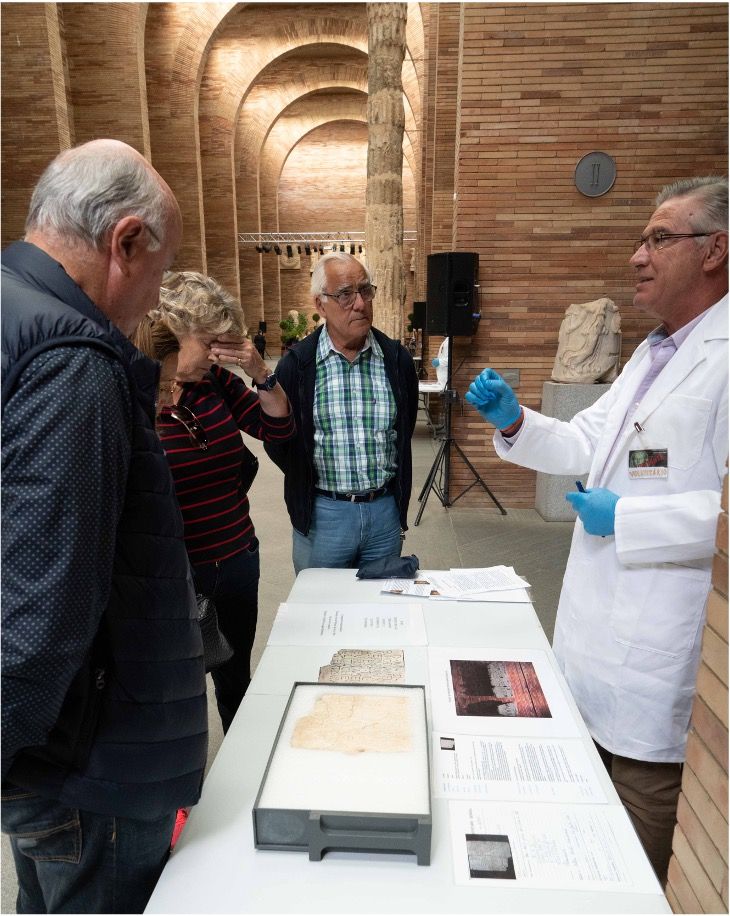 Un hombre con bata blanca y guantes azules explica a tres personas los objetos que se encuentran sobre una mesa en una sala de exposiciones. La sala tiene paredes de ladrillo, arcos y antiguas estatuas de piedra. Entre los objetos se encuentran tablillas y documentos expuestos bajo un cristal.