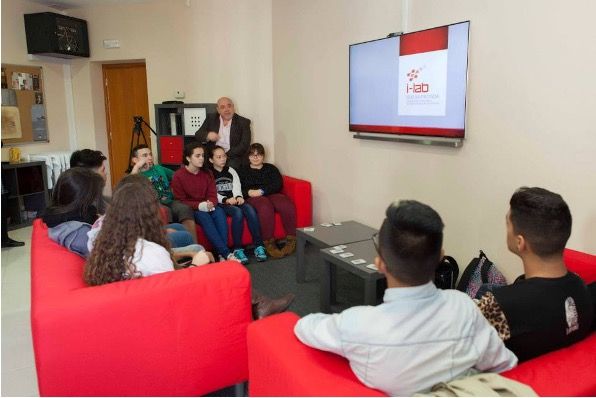 Un grupo de personas se sienta en sofás rojos en un ambiente que parece una sala de estar y escucha la presentación de un hombre mayor. El hombre está de pie cerca de una pantalla de televisión que muestra un logotipo con el texto "i-Lab". El público parece estar atento a la presentación.
