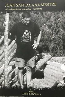 Fotografía en blanco y negro de un hombre de pie al aire libre junto a vigas de madera y una estructura parcialmente construida. Lleva gafas, camiseta negra y pantalones cortos. El texto en la parte superior dice: "Joan Santacana Mestre, profesor de Al savi, arqueólogo y museólogo.