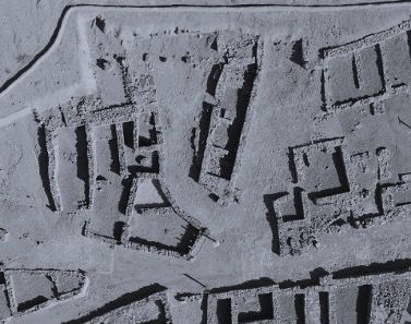 Imagen aérea en escala de grises que muestra las ruinas de un antiguo asentamiento. La imagen revela los contornos de estructuras rectangulares y cuadradas, probablemente restos de edificios, conectadas por estrechos caminos. El terreno parece plano con ligeras diferencias de elevación.