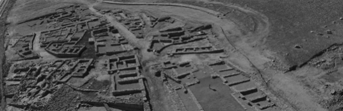 Black-and-white aerial view of ancient ruins, showing the remains of buildings and pathways laid out in a rectangular pattern within an arid landscape. The site appears to be an archaeological excavation, with partially intact walls and foundations visible.