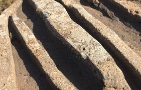 Primer plano de antiguas ranuras paralelas talladas en una superficie rocosa. Las ranuras están espaciadas uniformemente y recorren la imagen a lo largo, llenas de sombras y luz solar que resaltan los contornos. El área circundante parece seca y polvorienta.