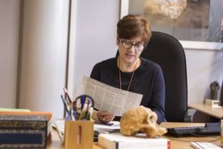 Una persona con cabello corto y castaño y gafas está sentada en un escritorio leyendo un periódico. En el escritorio hay varios útiles de oficina, libros, un teclado de computadora y un modelo de calavera. El fondo incluye una gran pintura en la pared.