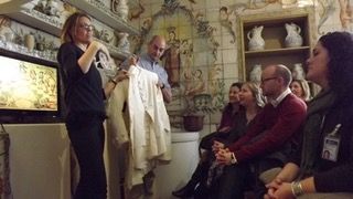 Un grupo de personas se sienta atentamente en una habitación decorada con estantes de cerámica y azulejos ornamentados en la pared, mientras una mujer y un hombre al frente sostienen una prenda blanca, posiblemente explicando o demostrando algo al respecto.