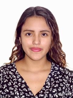 Una joven de cabello castaño ondulado, con una blusa negra con estampados florales blancos, está de pie sobre un fondo blanco liso. Tiene una expresión tranquila y mira directamente a la cámara.
