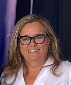 Una mujer de pelo largo y rubio con gafas sonríe a la cámara. Lleva una blusa blanca y pendientes de color turquesa. El fondo es oscuro con un degradado morado y negro.