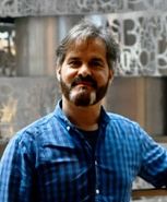 Un hombre con barba y bigote, que viste una camisa a cuadros azules, sonríe y está de pie en un interior. El fondo muestra una artística pared de metal con patrones abstractos.
