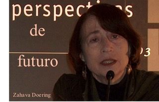 Una persona con cabello corto y oscuro habla por un micrófono. En el fondo se ve parcialmente el texto "Perspectivas de futuro". También se ve el nombre "Zahava Doering". La iluminación es tenue y se centra en el rostro de la persona.