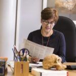 Una persona con cabello corto y castaño y gafas está sentada en un escritorio leyendo un periódico. En el escritorio hay varios útiles de oficina, libros, un teclado de computadora y un modelo de calavera. El fondo incluye una gran pintura en la pared.