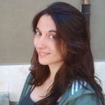Elena Asensio. Una mujer con el pelo largo y oscuro y vestida con una camisa gris y una chaqueta verde claro está al aire libre. Ella sonríe levemente y mira hacia la cámara. El fondo presenta una pared beige y parte de la estructura de un edificio.