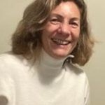 Elena Pol. Una mujer con el pelo hasta los hombros y un jersey de cuello alto de color claro sonríe cálidamente a la cámara. Está en el interior, con una pared de color claro y cuadros enmarcados parcialmente visibles al fondo.