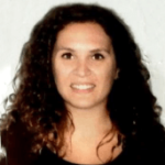 Sabrina Armenio. Una mujer con cabello largo, rizado y oscuro sonríe a la cámara. Lleva una blusa oscura y está colocada sobre un fondo liso y de color claro.