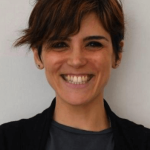 Yone Castillo. Una mujer de pelo corto y castaño y que viste una chaqueta oscura sonríe ampliamente a la cámara. El fondo es de color claro y liso.