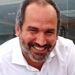 Abel Ponce. Un hombre de pelo corto oscuro, barba y bigote sonríe. Viste una camisa blanca y está fotografiado sobre un fondo con paneles de vidrio y un indicio de estructura de madera.