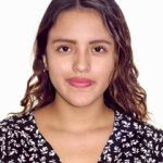 Una joven de cabello castaño ondulado, con una blusa negra con estampados florales blancos, está de pie sobre un fondo blanco liso. Tiene una expresión tranquila y mira directamente a la cámara.