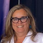 Una mujer de pelo largo y rubio con gafas sonríe a la cámara. Lleva una blusa blanca y pendientes de color turquesa. El fondo es oscuro con un degradado morado y negro.