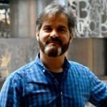 Un hombre con barba y bigote, que viste una camisa a cuadros azules, sonríe y está de pie en un interior. El fondo muestra una artística pared de metal con patrones abstractos.