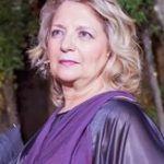 Pilar Caldera. Una mujer con cabello rubio hasta los hombros está parada al aire libre. Lleva un traje morado y negro y mira ligeramente hacia su derecha. El fondo está desenfocado, con vegetación y árboles.