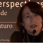 Una persona con cabello corto y oscuro habla por un micrófono. En el fondo se ve parcialmente el texto "Perspectivas de futuro". También se ve el nombre "Zahava Doering". La iluminación es tenue y se centra en el rostro de la persona.